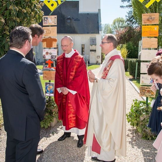 Zwei Priester in roten und weißen Gewändern unterhalten sich vor einer Kirche, mit einem Banner, das 'BARM HERZIT' darüber steht.