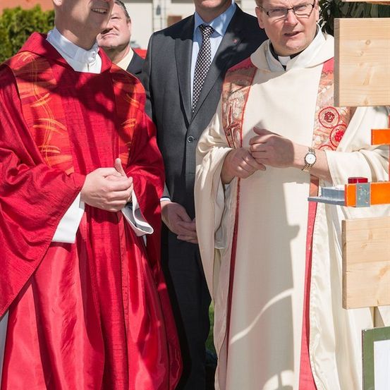 Drei Männer in formeller Kleidung, zwei in geistlichen Gewändern, einer in einem Anzug, stehen draußen an einem sonnigen Tag.