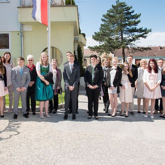 Eine Gruppe von Menschen in formeller Kleidung, möglicherweise für einen Abschluss, steht vor einem Gebäude mit einer französischen Flagge.