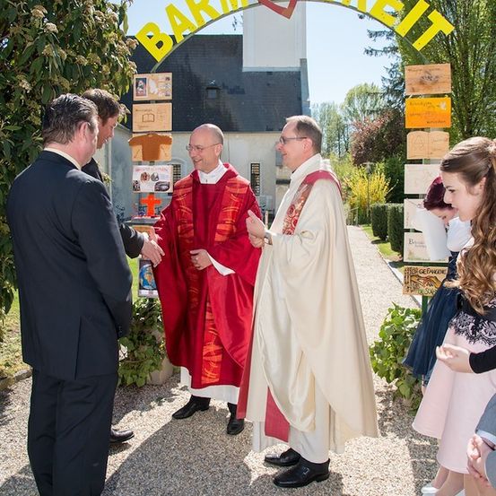 Zwei Priester in traditionellen Gewändern begrüßen Gäste am Eingang einer Kirche. Der Bogen über ihnen trägt den deutschen Schriftzug 'Barmherzigkeit'.