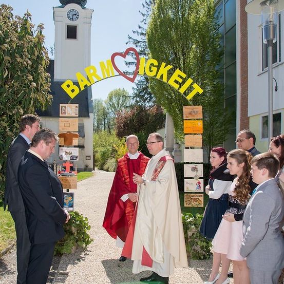 Eine Gruppe von Menschen, einschließlich Priester, steht vor dem Eingang einer Kirche mit einem Schild, das 'Barmherzigkeit' besagt. Die Umgebung ist draußen mit einem klaren Himmel und einem Uhrturm im Hintergrund sichtbar.