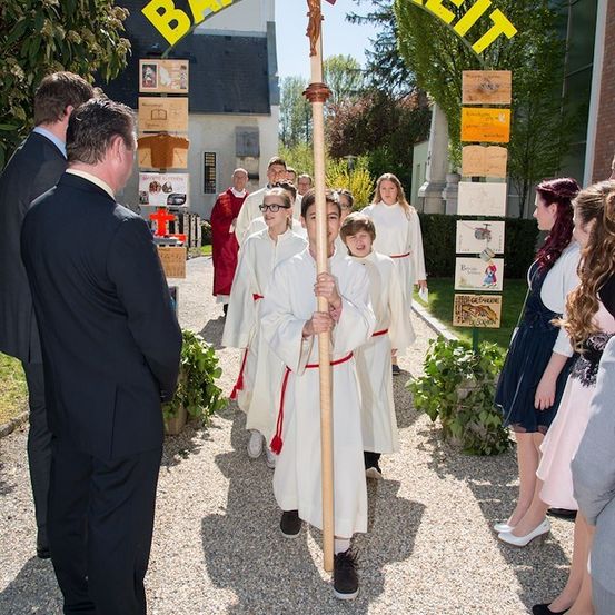 Eine Gruppe von Kindern in weißen Roben und mit Brillen hält ein Kreuz und nimmt an einer Prozession teil. Sie werden von Erwachsenen begleitet, einige in formeller Kleidung. Die Szene spielt sich in einem Garten mit einem Gebäude und Pflanzen im Hintergrund ab.