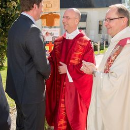 Drei Männer in formeller Kleidung, zwei in Rot und einer in Grau, stehen vor einer Kirche, lächeln und unterhalten sich. Hinter ihnen ist ein Gebäude mit einem Kreuz und einem Schild zu sehen.