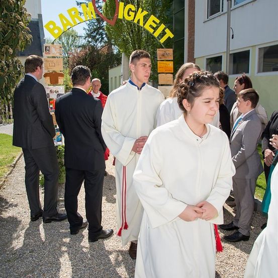 Eine Gruppe von Menschen in weißen Gewändern steht vor einem Gebäude mit einem Schild, das 'Barmherzigkeit' besagt. Die Menschen schauen nach unten und scheinen zu beten.