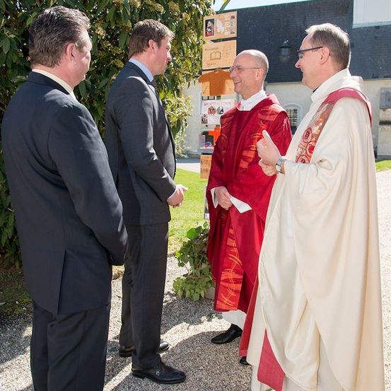 Vier Männer in formeller Kleidung, zwei in schwarzen Anzügen und zwei in kirchlichen Gewändern, unterhalten sich vor einer Kirche.