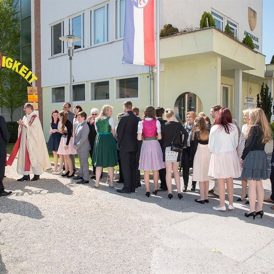 Eine Gruppe von Menschen in formeller Kleidung steht in einer Reihe vor einem Gebäude mit einem Banner, das 'IGKEIT' besagt. Ein Priester führt die Gruppe an.