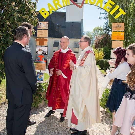 Zwei Priester in traditionellen Gewändern stehen unter einem Schild mit der Aufschrift 'Barmherzigkeit' und einem Herzsymbol, im Gespräch mit einer Gruppe von Menschen vor einer Kirche.