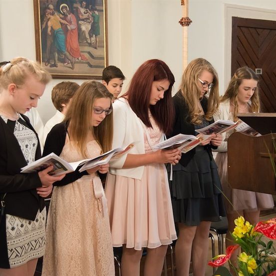 Eine Gruppe junger Frauen in verschiedenen Outfits steht in einer Kirche und liest aus einem Buch.