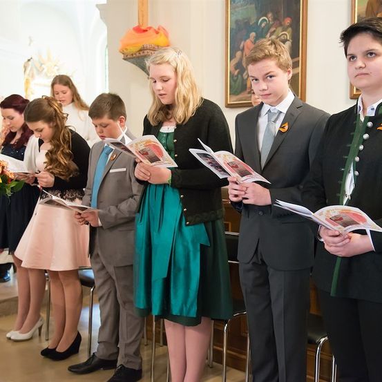 Eine Gruppe junger Menschen in formeller Kleidung steht in einer Kirche, hält Bücher und schaut nach unten.