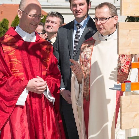 Zwei Priester in prächtigen Gewändern unterhalten sich mit einem Mann in einem Anzug. Hinter ihnen ist ein hölzernes Bauwerk teilweise zu sehen.