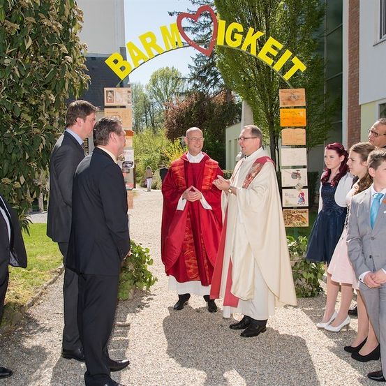 Zwei Priester in roten und weißen Gewändern unterhalten sich mit einer Gruppe von Menschen, möglicherweise einer Familie, vor einem Gebäude mit einem Schild, das 'Barmherzigkeit' besagt.