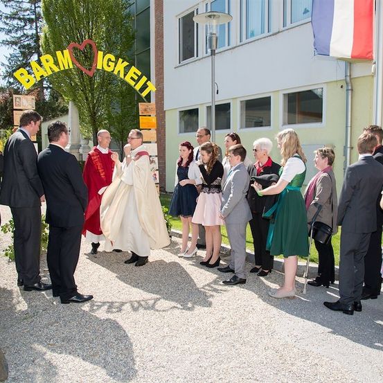 Eine Gruppe von Menschen in formeller Kleidung steht vor einem Gebäude mit einem Schild, das 'Barmherzigkeit' besagt. Ein Priester spricht zu einem Paar in der Mitte der Gruppe.