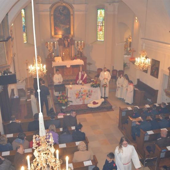 Ein Kircheninneres mit einem Priester am Altar, umgeben von Kerzen und Blumen, mit einer Gemeinde, die auf Kirchenbänken sitzt.