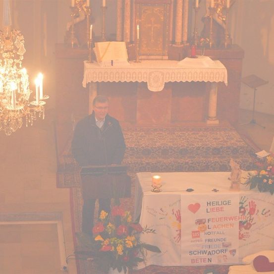 Ein Mann steht an einem Podest in einer Kirche, mit einer Kerze und Blumen vor ihm. Dahinter befindet sich ein Altar mit Kerzen und einem Schild, das 'Heilige Liebe' besagt.