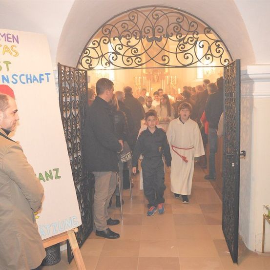 Eine Gruppe von Menschen betritt eine Kirche, mit einem Banner auf der linken Seite und einem kunstvollen Tor auf der rechten Seite.