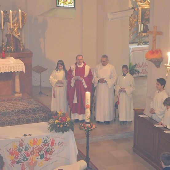 Eine Gruppe von Menschen in weißen Gewändern steht in einer Kirche, hält Kerzen und liest aus einem Buch. Ein Priester in einem rot-weißen Gewand steht vor ihnen.