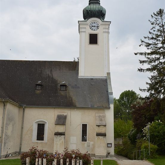 Eine kleine Kirche mit einem hohen Turm und einer Uhr, umgeben von Grün und einem Kiesweg.