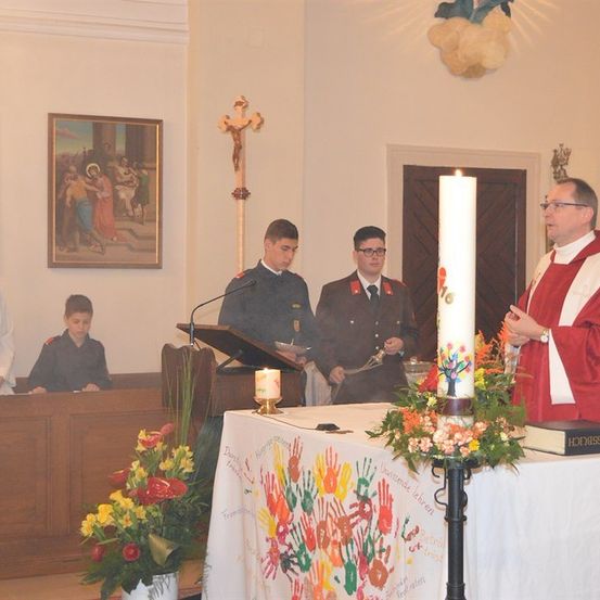 Ein Priester steht mit einer Kerze am Altar, flankiert von zwei Männern in Uniform, während ein junger Junge und eine Frau im Hintergrund beobachten. Der Altar ist mit Blumen und einem Buch geschmückt.