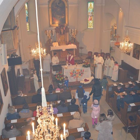 Ein Kircheninneres mit einem Priester am Altar, umgeben von Kerzen, Blumen und Besuchern auf den Kirchenbänken. Der Altar ist mit einem weißen Tuch und einer Blumenarrangement geschmückt. Die Kirche verfügt über Buntglasfenster, Gemälde und Kronleuchter.