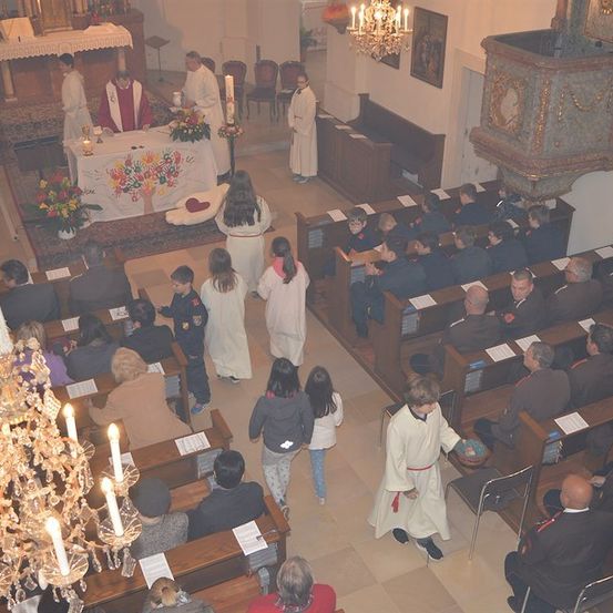 Eine religiöse Zeremonie findet in einer Kirche statt, mit einem Priester am Altar, einer Gemeinde in den Bänken und einem Chor im Hintergrund. Der Altar ist mit Blumen und Kerzen geschmückt.