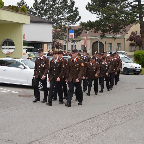 Eine Gruppe uniformierter Personen, möglicherweise Militärangehörige, marschiert bei einer Parade auf einer Straße. Sie tragen Hüte, Medaillen und Schuhe. Es sind Autos am Straßenrand geparkt.