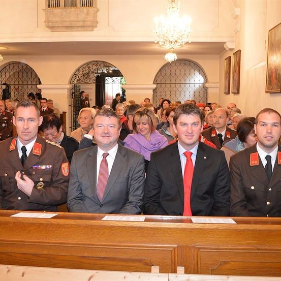 Vier Männer in formeller Kleidung, möglicherweise Militäroffiziere, sitzen in der vorderen Reihe einer Kirche. Hinter ihnen sitzen eine Menge Menschen auf den Kirchenbänken. Der Raum ist gut beleuchtet mit Kronleuchtern und Gemälden an der Wand.