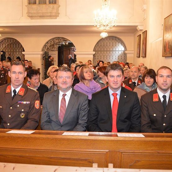 Vier Männer in formeller Kleidung, zwei in Militäruniformen und zwei in Anzügen, sitzen in der vordersten Reihe einer Kirche. Hinter ihnen sitzen eine Menge Menschen in verschiedenen Outfits auf Kirchenbänken. Der Raum ist gut beleuchtet mit Kronleuchtern und gerahmten Bildern an der Wand.