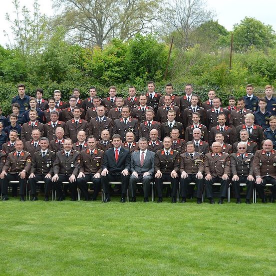 Eine Gruppe uniformierter Personen, möglicherweise Militäroffiziere, posiert für ein Gruppenfoto auf einer Rasenfläche mit Grünpflanzen im Hintergrund.