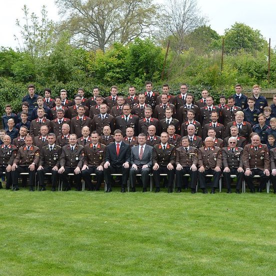 Eine Gruppe uniformierter Personen, möglicherweise Militärangehörige, posiert für ein Foto auf einem gepflegten Rasen mit Grünpflanzen im Hintergrund.
