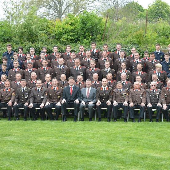 Eine Gruppe uniformierter Personen, darunter Männer und Frauen, sitzt auf Bänken im Freien. Sie tragen Militäruniformen und einige haben Medaillen. Hinter ihnen stehen mehrere Bäume.