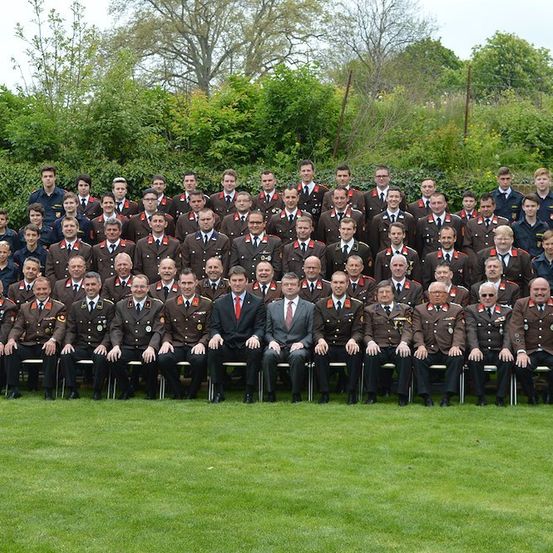 Eine große Gruppe von Militärangehörigen in Uniform, einige sitzend und einige stehend, posiert für ein Gruppenfoto auf einer Rasenfläche mit Bäumen im Hintergrund.