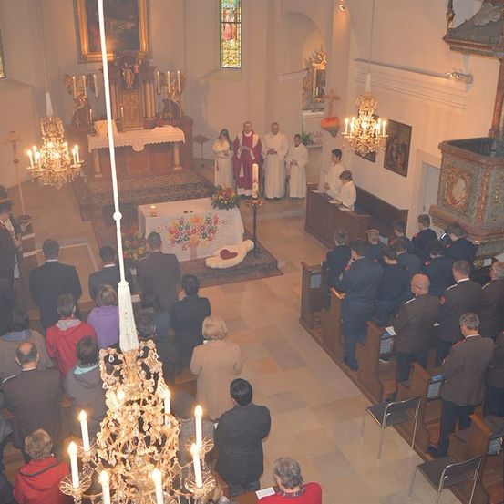 Eine Gruppe von Menschen ist in einer Kirche für eine religiöse Zeremonie versammelt. Der Altar ist mit Blumen geschmückt, und Priester stehen davor. Kerzen und Kronleuchter erhellen den Raum.