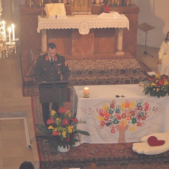 Ein Militäroffizier steht in einer Kirche an einem Rednerpult, vor ihm ein Tisch mit einer weißen Tischdecke und einem Baum aus Handabdrücken. Auf dem Tisch brennt eine Kerze, und eine Kristallkronleuchter hängt von der Decke. Eine Frau sitzt auf der rechten Seite.