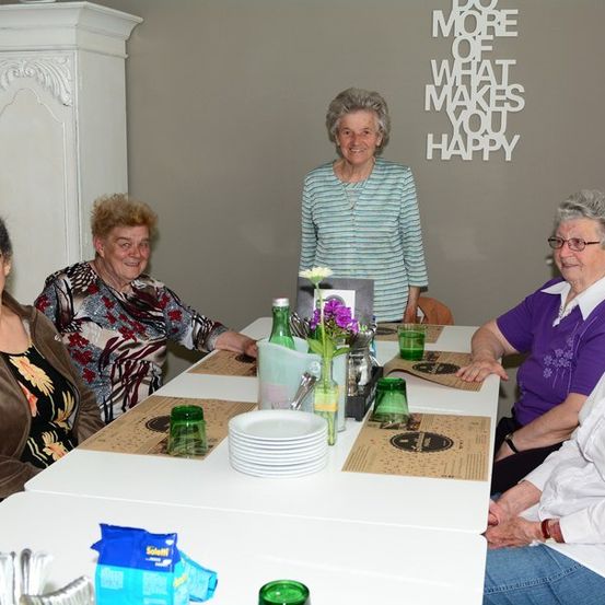Fünf Frauen sitzen um einen Tisch in einem Raum. Eine Frau steht hinter ihnen. Der Tisch hat Teller, Gläser und Blumen. Die Wand hinter ihnen hat einen motivierenden Spruch.