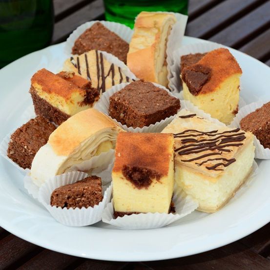 Ein weißer Teller mit einer Auswahl an Desserts, darunter Käsekuchen, Brownies und andere Gebäckstücke, die ordentlich in Papierbechern auf einem Holztisch angeordnet sind.