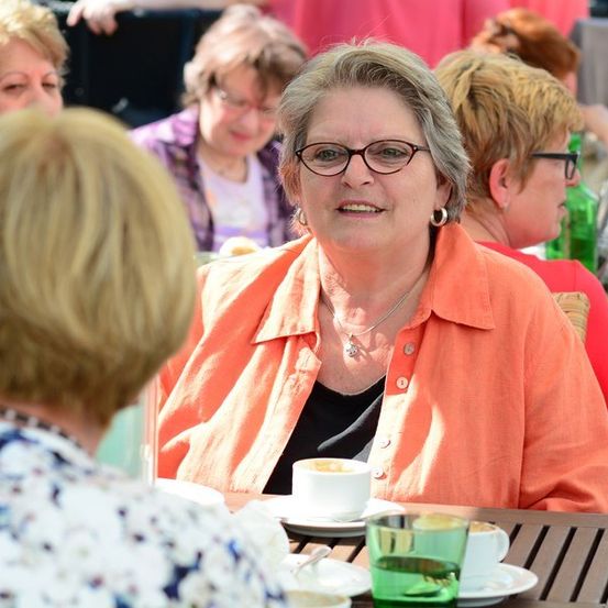 Eine Gruppe von Frauen in einem Café im Freien, eine Frau in einem orangefarbenen Shirt lächelt und schaut eine andere Frau an.