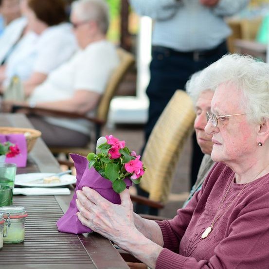Eine ältere Frau hält einen Strauß rosa Blumen und sitzt mit anderen älteren Menschen an einem Tisch in einem Garten.