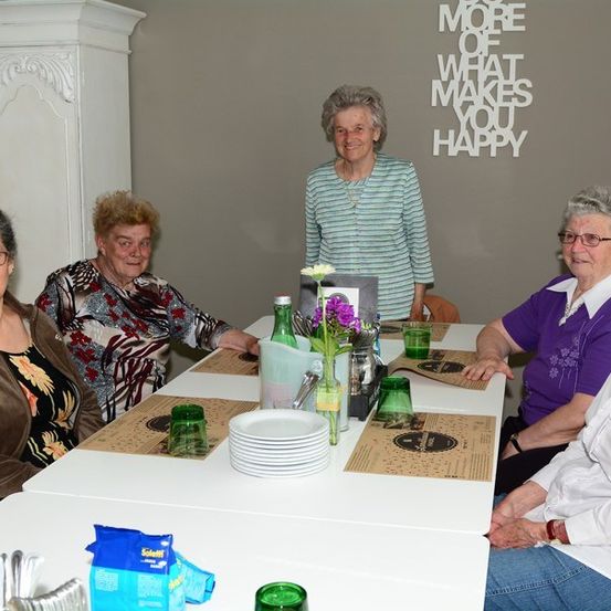 Eine Gruppe älterer Frauen sitzt um einen Tisch und lächelt für ein Foto. Der Tisch ist mit Tellern, Gläsern und einer Blumenarrangement gedeckt. Hinter ihnen ist eine Wand mit dem Text 'MEHR VON DEM, WAS DICH GLÜCKLICH MACHT.'