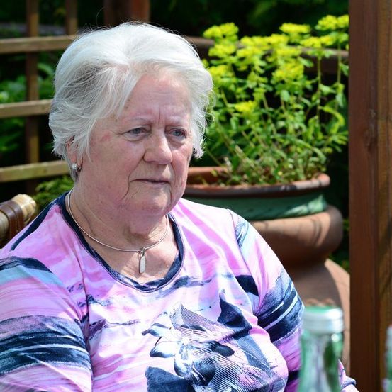 Eine ältere Frau mit weißen Haaren sitzt in einem Gartenstuhl und trägt ein rosa T-Shirt mit Blumenmuster. Hinter ihr steht eine Topfpflanze mit gelben Blüten.