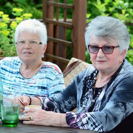 Zwei ältere Frauen sitzen an einem Tisch im Garten, eine hält eine Tasse und die andere eine Flasche mit grüner Flüssigkeit. Sie tragen beide eine Brille und Schmuck.