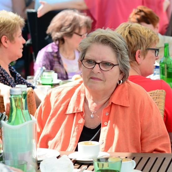 Eine ältere Frau mit Brille sitzt in einem Café im Freien, umgeben von anderen Frauen. Vor ihr steht eine Tasse Kaffee und eine Flasche Limonade.