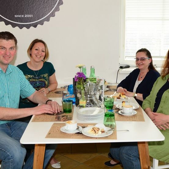 Vier Personen sitzen an einem Tisch mit Tellern, Tassen und einer Blumenvase. Der Mann links trägt ein kariertes Hemd und die Frau rechts ein grünes Kleid. Hinter ihnen befindet sich eine Wand mit Logo und Fenster.