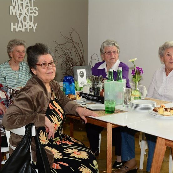 Fünf ältere Frauen sitzen um einen Tisch herum, möglicherweise bei einem gesellschaftlichen Treffen. Der Tisch ist mit Tellern, einer Blumenvase und einer Flasche gedeckt. Die Frau in der Mitte trägt eine Brille und ein Blumenkleid. Hinter ihnen zeigt eine Wand den Text 'Was macht dich glücklich?'