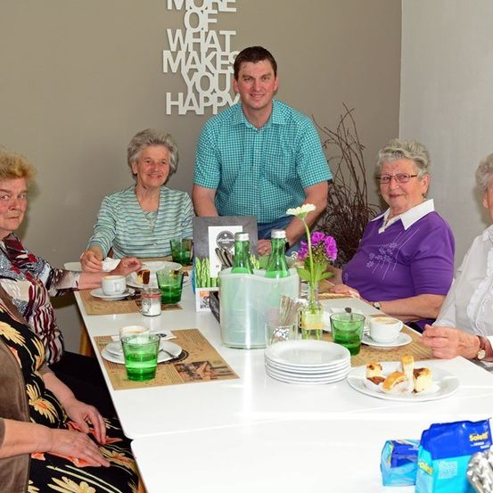 Eine Gruppe älterer Frauen und ein Mann sitzen um einen Tisch mit Tellern, Tassen und Blumen. Sie lächeln und scheinen ihre Mahlzeit zu genießen. Der Mann steht hinter ihnen und es gibt eine Wand mit einem Zitat dahinter.