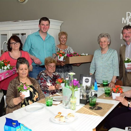 Eine Gruppe von Menschen, sowohl Männer als auch Frauen, versammelt sich um einen Tisch, hält Blumentöpfe und lächelt für ein Foto. Der Tisch ist mit Tellern, Gläsern und einer Vase mit Blumen gedeckt. Ein Schild an der Wand steht 'Mach mehr'.
