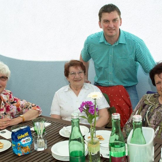 Eine Gruppe von vier Personen, drei Frauen und ein Mann, sitzen an einem Tisch mit Tellern, Blumen und Flaschen. Sie lächeln und posieren für ein Foto.