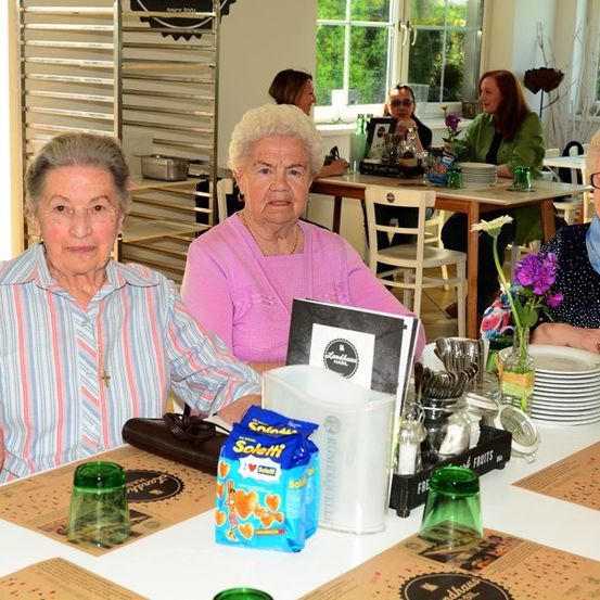 Drei ältere Frauen sitzen an einem Tisch in einem Restaurant, lächeln und posieren für ein Foto. Der Tisch ist mit Tellern, Gläsern und einer Speisekarte gedeckt. Hinter ihnen sitzen andere Personen an Tischen, und der Raum ist mit natürlichem Licht gefüllt.