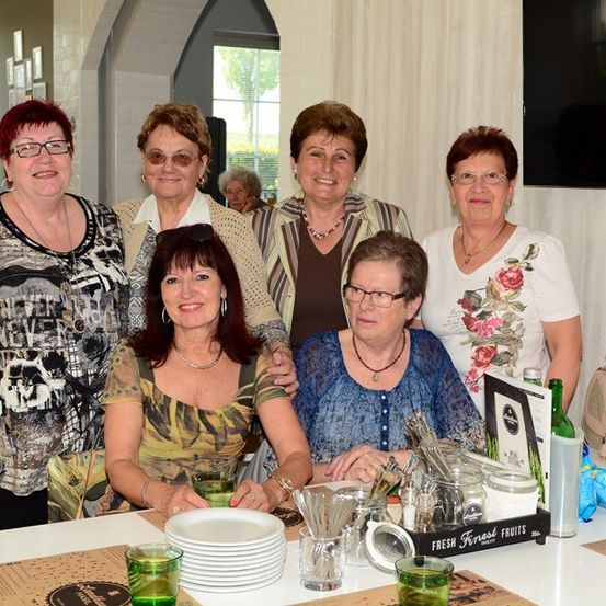 Sechs Frauen versammeln sich um einen Tisch, lächeln und posieren für ein Foto. Sie tragen Brillen, Halsketten und Ohrringe. Der Tisch hat Teller, Gläser und eine Schachtel frische Früchte. Hinter ihnen ist ein Fenster mit Blick auf einen Garten.
