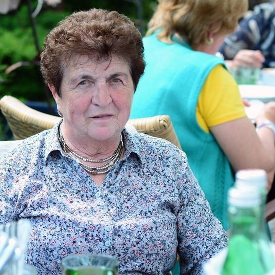 Eine ältere Frau mit kurzen, lockigen Haaren sitzt am Tisch, trägt ein Blumenhemd und mehrere Halsketten. Hinter ihr isst eine andere Frau in einer blauen Weste.