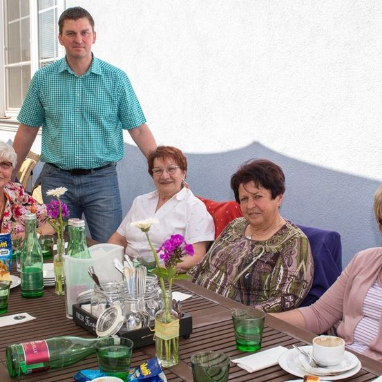 Eine Gruppe von Frauen und einem Mann sitzen um einen Tisch und posieren für ein Foto. Der Tisch ist mit Tellern, Gläsern und Blumen gedeckt. Der Mann steht hinter den Frauen, und sie sind vor einer weißen Wand.